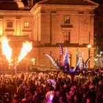 Public square at night filled with celebrating people illuminated by giant art and flaming sculpture