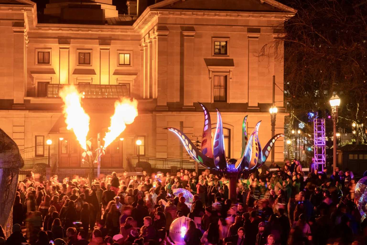 Public square at night filled with celebrating people illuminated by giant art and flaming sculpture