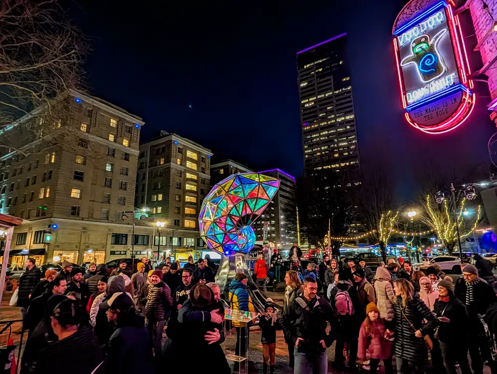 A crowd around a giant glowing nautilus shell art installation in a busy city square
