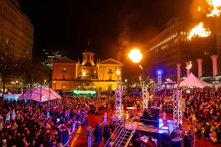 Giant crowd surrounds stage in city square illuminated by art and a flaming piano performance
