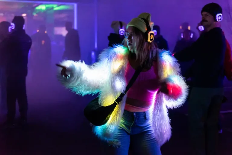 Woman wearing white furry LED illuminated coat and glowing headphones dances and points in a purple glowing foggy outdoor space at night