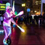 Man in glowing white tuxedo juggles glowing bowling pins while riding a one wheel for an adoring crowd
