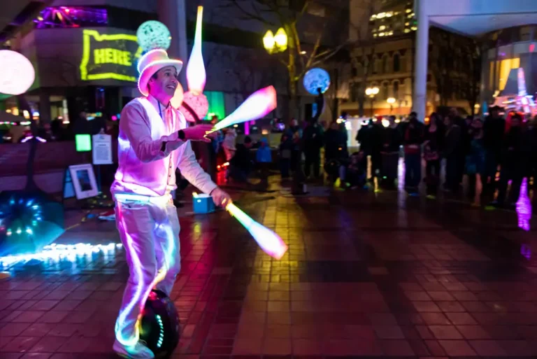 Man in glowing white tuxedo juggles glowing bowling pins while riding a one wheel for an adoring crowd