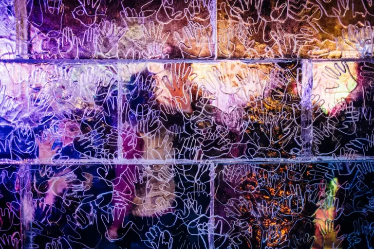 Clear ice-sculpture wall covered with hands engraved into the purple and yellow glowing ice at night.