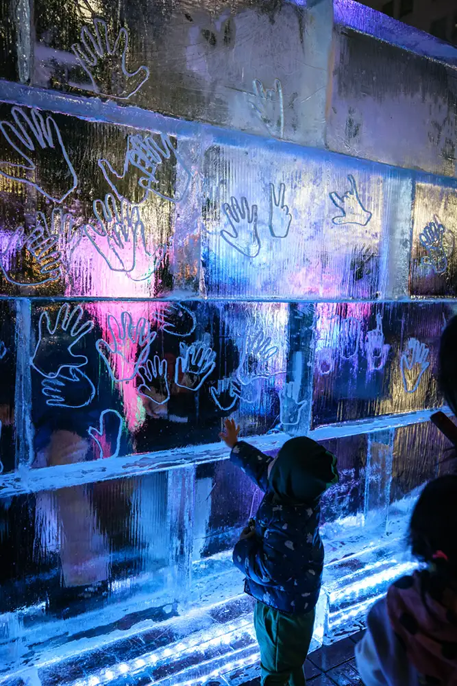 Small child dressed for winter places hand on clear ice-sculpture wall with hands engraved into the blue glowing ice at night.