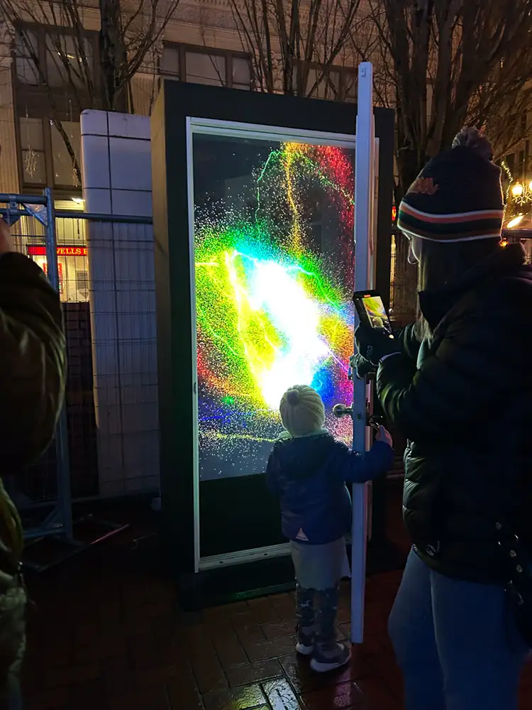 Small child dressed for winter opens door to art sculpture which presents a screen of glowing cosmic lights at night