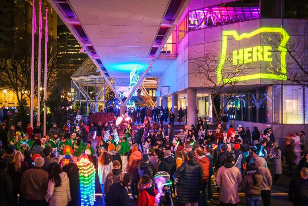 Crowded downtown city people celebrating light festival at night glowing yellow sign reads "HERE" in Oregon State shaped frame.
