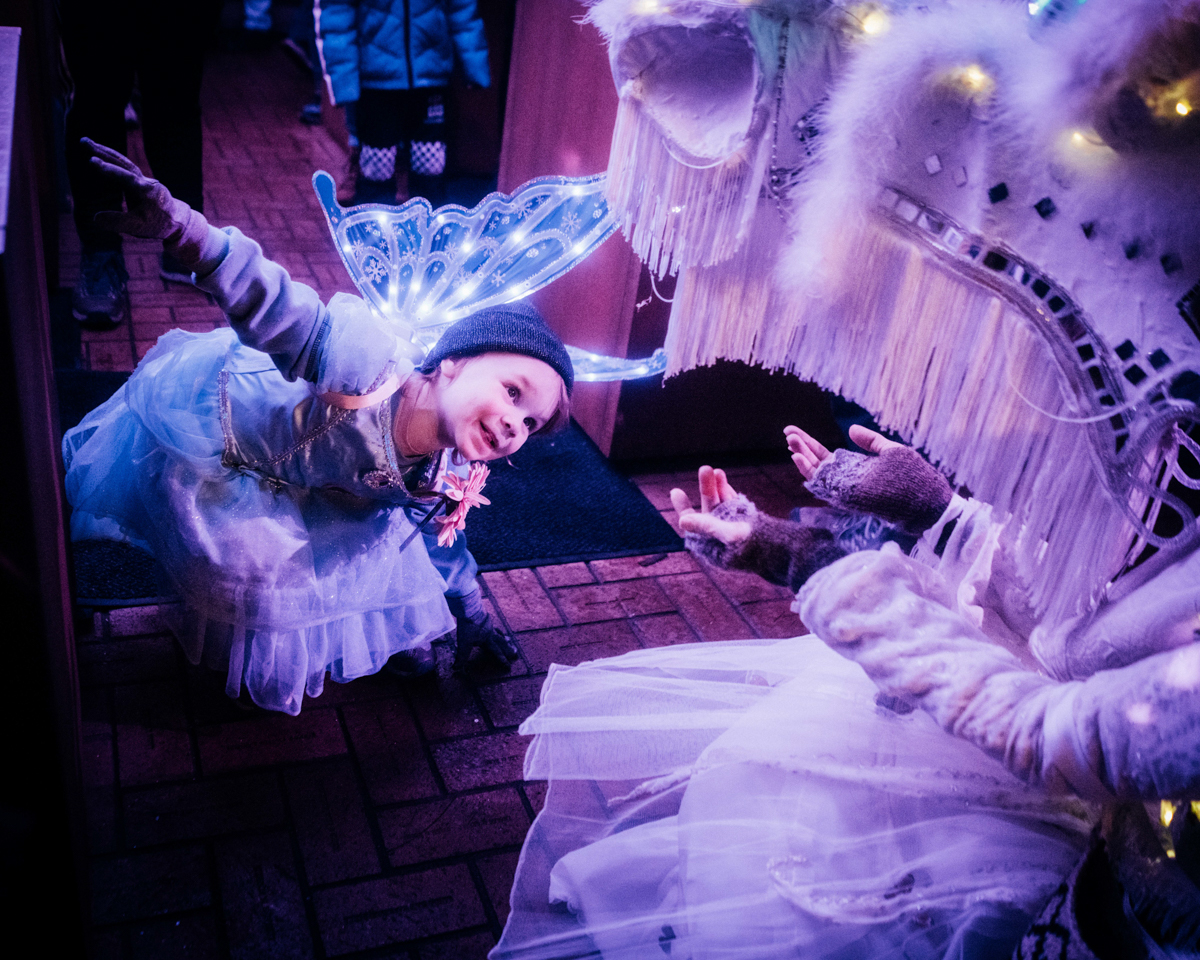 Little girl wearing illuminated butterfly wings peers up under a costumed dragon