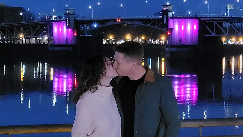 couple kisses at twilight with illuminated bridge behind them