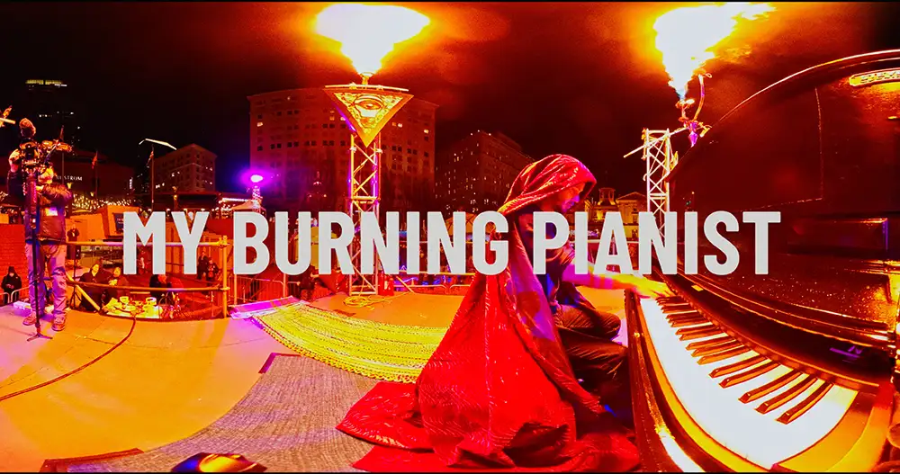 screenshot of piano player wearing red hooded cape playing while propane fire shoots straight up