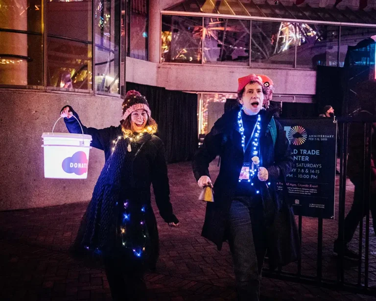 two woman excitedly dance outdoors at night holding 'donate' buckets