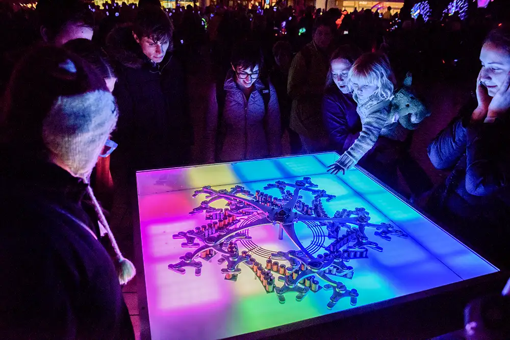 people surround glowing interactive art table outdoors at night