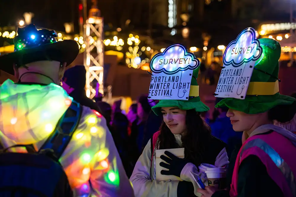 Two teens wearing illuminated hats reading "Survey" speak to a Festival fan outdoors at night