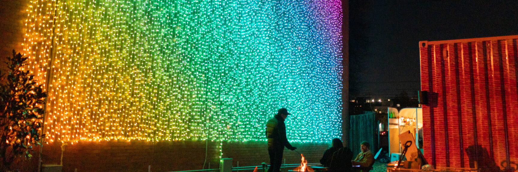 Giant wall illuminated in colorful rainbow while people enjoy a campfire at night